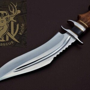 May include: A silver hunting knife with a brown wooden handle and a serrated edge. The knife has a brass accent on the handle and a black accent on the blade. The knife is on a brown background. The text "Wild Deer Knife Damascus" is visible in the upper left corner of the image.