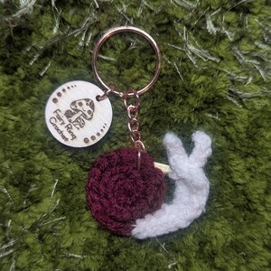 May include: A handmade keychain featuring a burgundy crocheted snail with a white body. The keychain includes a wooden tag with the text "Fairy Ring Crochet" and a mushroom graphic. The keychain is attached to a copper-colored ring.