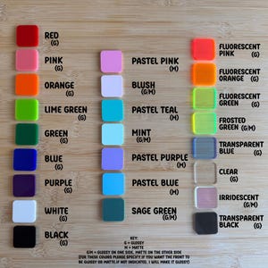 May include: A wooden surface displays a color chart of square acrylic pieces. Colors include red, pink, orange, lime green, green, blue, purple, white, and black, with fluorescent and pastel shades. The key indicates glossy or matte finishes.