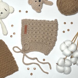May include: A tan crocheted baby bonnet with a textured pattern and a small brown leather tag. The bonnet has tie strings and is surrounded by other crocheted items, buttons, and cotton stems.
