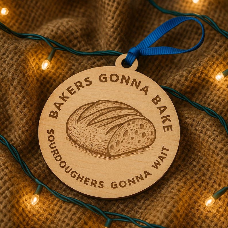 May include: A round wooden ornament with the text "BAKERS GONNA BAKE SOURDOUGHERS GONNA WAIT" encircling a detailed illustration of a loaf of bread. A blue ribbon is attached for hanging. The ornament is set against a brown, textured background.