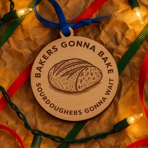 May include: Wooden Christmas ornament with a bread loaf illustration and the text "SOURDOUGHERS GONNA BAKE, GONNA WAIT". The ornament has a blue ribbon for hanging and is surrounded by red and green ribbons and Christmas lights.