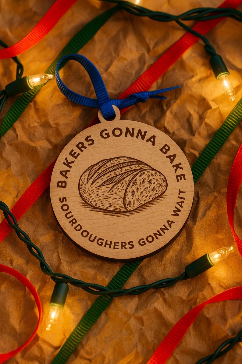May include: Wooden Christmas ornament with a bread loaf illustration and the text "SOURDOUGHERS GONNA BAKE, GONNA WAIT". The ornament has a blue ribbon for hanging and is surrounded by red and green ribbons and Christmas lights.