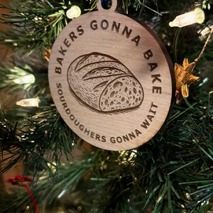 May include: Wooden Christmas ornament with a blue ribbon, featuring the text "Bakers Gonna Bake, Sourdoughers Gonna Wait" around a detailed illustration of a loaf of bread. The ornament is hanging on a Christmas tree.