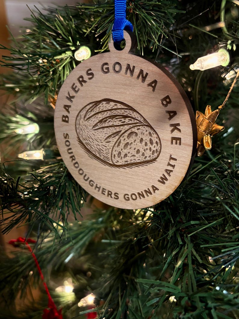 May include: Wooden Christmas ornament with a blue ribbon, featuring the text "Bakers Gonna Bake, Sourdoughers Gonna Wait" around a detailed illustration of a loaf of bread. The ornament is hanging on a Christmas tree.