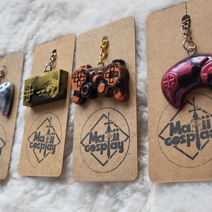 May include: Four different coloured video game controller keychains on brown card stock with the text "Maili Cosplay" printed on each card. The keychains are silver, gold, black and red.