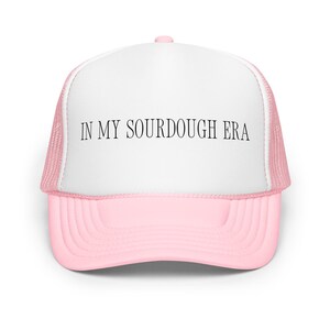 May include: Pink and white trucker hat with the text "IN MY SOURDOUGH ERA" in black. The hat has a pink brim and a white front panel with the text. The mesh backing is pink.