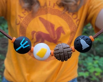 May include: A brown leather cord necklace with five decorative beads. The beads are black, white, and brown, with symbols including a trident, a building, and a yellow shape. The necklace is worn over an orange t-shirt.