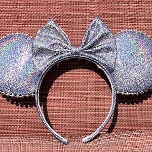 Può includere: Orecchie di Minnie Mouse con glitter argentato e un fiocco argentato. Le orecchie hanno un bordo di perline.