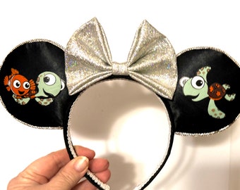 May include: Black mouse ears with a silver glitter bow. The ears feature a green sea turtle and an orange and white clownfish.