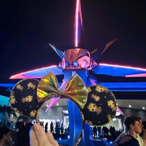 May include: A pair of black and gold Minnie Mouse ears with a gold bow and a pattern of Groot faces on the ears. The ears are in front of a large, colorful, futuristic structure with pink and blue lights.