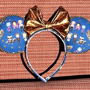 May include: A pair of blue and gold mouse ears with a gold bow. The ears are decorated with characters from the Guardians of the Galaxy movie. The text "Guardians of the Galaxy" is printed on the ears.
