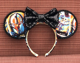 May include: Black and white mouse ears with a black sequined bow. The ears feature images of the Mandalorian and "The Child" from the Star Wars series. The left ear has the text "MANDO" and the right ear has the text "THE CHILD".