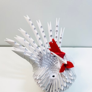 May include: White origami rooster with red accents. The rooster has a large, elaborate tail and a red comb and wattle.
