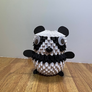 May include: A black and white origami panda with googly eyes. The panda is made of folded paper and has a black nose and ears.