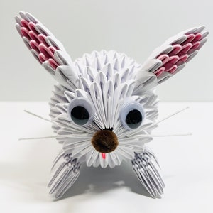 May include: White origami rabbit with pink ears, black eyes, and a brown felt nose. The rabbit is sitting on a white surface.