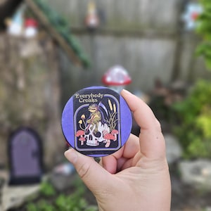 May include: A round, purple coaster with the text "Everybody Croaks" and a graphic of a frog grim reaper on a skull, surrounded by mushrooms and reeds. The coaster is held in a hand against a blurred outdoor background.