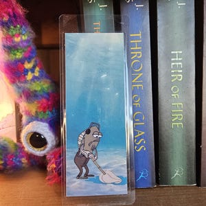 May include: A rectangular, clear-laminated bookmark featuring a printed illustration of a grey fish character with headphones, mopping a blue underwater floor. The bookmark is positioned vertically in front of several dark-spined hardcover books.