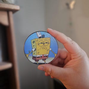 May include: A round wooden coaster featuring a grumpy cartoon character, SpongeBob SquarePants, with a blue background. The coaster has a black border and is held in a hand.