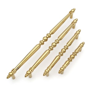 May include: Set of four gold-colored cabinet pulls with ornate detailing. The pulls feature a combination of straight bars and decorative, beaded sections. The hardware is designed for furniture and cabinetry, offering a classic aesthetic.