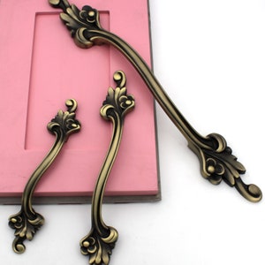 May include: Three ornate, antique gold-toned cabinet door pulls with a decorative floral design. The pulls are arranged in a row on a pink surface.