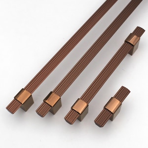 May include: Four bronze-coloured cabinet pulls and knobs. The pulls have a textured, linear design with a rectangular base and a copper-coloured accent. The knobs have a similar design, but are shorter. These hardware pieces are ideal for kitchen or bathroom cabinets.