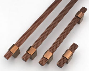 May include: Four bronze-colored cabinet pulls and knobs. The pulls have a textured, linear design with a rectangular base and a copper-colored accent. The knobs have a similar design, but are shorter. These hardware pieces are ideal for kitchen or bathroom cabinets.