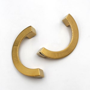 Könnte beinhalten: Zwei goldfarbene Metall-Schrankgriffe mit einer gebogenen Form. Die Griffe haben ein quadratisches Ende.