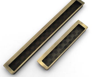 May include: Two rectangular bronze-colored recessed cabinet pulls. The pulls have a dark, checkered pattern in the recessed area. One pull is larger than the other. The pulls are set against a white background.