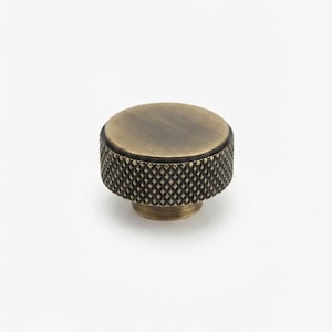 May include: A round cabinet knob with an antique brass finish. The top surface is smooth and flat, while the cylindrical side features a textured, diamond-patterned knurled grip. The hardware has a metallic, darkened gold-toned appearance.