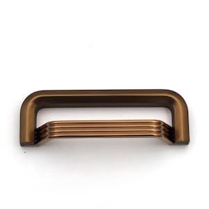 Può includere: Una maniglia per mobili rettangolare con una finitura in bronzo opaco e liscia. La maniglia presenta una cornice esterna solida con una parte superiore arrotondata e una sezione centrale composta da tre barre orizzontali, rialzate e scanalate.