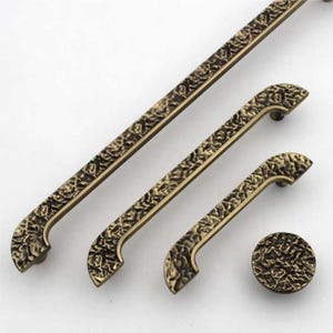 Può includere: Un set di ferramenta per mobili composto da tre maniglie per cassetti in metallo allungate e un pomello circolare. Ogni pezzo ha una finitura in bronzo scuro con una superficie testurizzata e irregolare che ricorda la pietra grezza.