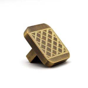 May include: A rectangular cabinet knob with a brushed antique brass finish. The front face features a recessed diamond lattice pattern with small, raised dots along the grid lines, providing a textured surface on the metallic hardware piece.