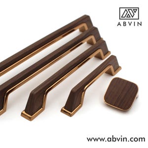 Könnte beinhalten: Ein Satz Holzschrankgriffe und ein Knopf mit goldfarbenem Besatz. Die Griffe sind rechteckig mit einer dunkelbraunen Holzmaserung. Der Knopf ist quadratisch. Der Markenname "ABVIN" befindet sich in der oberen rechten Ecke, und die Website steht unten.