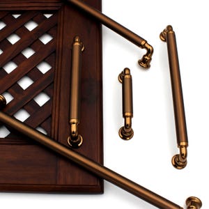 Könnte beinhalten: Ein Satz aus fünf gebürsteten Bronze-Schrankgriffen. Die Griffe sind in verschiedenen Größen und Stilen erhältlich, darunter ein langer Stangen-Griff, ein kurzer Stangen-Griff und zwei kleinere Griffe mit einem geschwungenen Design. Die Griffe werden vor einem dunklen Holzhintergrund gezeigt.