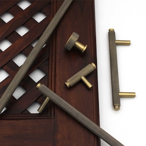 May include: An assortment of antique brass cabinet hardware, including handles and knobs. The hardware features a textured, diamond-patterned grip and a brass finish. The pieces are arranged on a dark wood surface, showcasing their design and style.