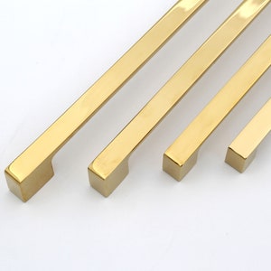 May include: Four gold-toned metal cabinet pulls of varying lengths. The pulls have a rectangular shape with rounded edges.