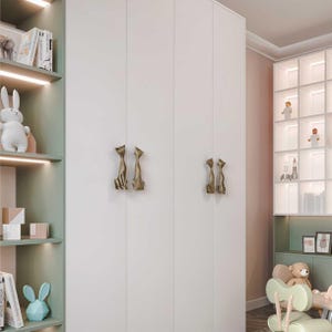 Pode incluir: Um guarda-roupa branco com puxadores dourados em forma de gato. Os puxadores estão posicionados em pares nas portas. O guarda-roupa faz parte de um quarto infantil, com prateleiras exibindo brinquedos e livros.