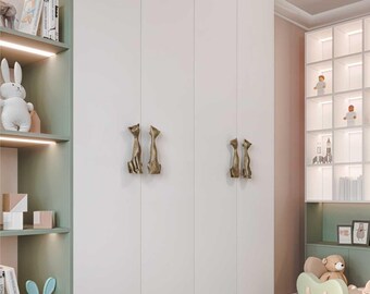 May include: A white wardrobe with brass cat-shaped handles. The handles are positioned in pairs on the doors. The wardrobe is part of a child's room, with shelves displaying toys and books. The room has a soft color palette.