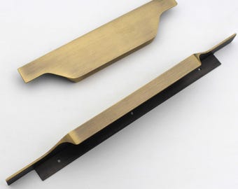 May include: Two brushed brass cabinet pulls with a long, flat, horizontal design. The handles feature a metallic gold finish with a subtle grain texture. The bottom edge of the longer handle shows a dark brown mounting plate with two screw holes.