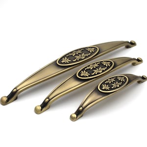 Puede incluir: Tres tiradores de armario de latón antiguo de diferentes tamaños. Cada tirador presenta un centro ovalado con un diseño floral. Los tiradores tienen forma curva con extremos redondeados, sobre fondo blanco.