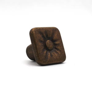 May include: A square cabinet knob with a dark bronze finish and a rounded, sunburst-like embossed pattern in the centre. The knob features a smooth, slightly weathered texture and a cylindrical base extending from the back of the square face.