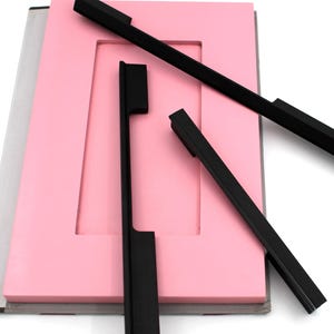 Könnte beinhalten: Vier schwarze Schrankgriffe auf einem rosa Hintergrund. Die Griffe sind rechteckig mit einer flachen, glatten Oberfläche.