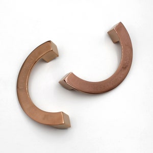 May include: Two rose gold cabinet pulls in a semi-circular design. Each pull has a square end and a curved body. The pulls are made of metal and are set against a white background. These are ideal for updating kitchen or bathroom cabinets.