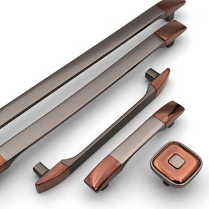 Könnte beinhalten: Ein Satz von fünf Schrankgriffen und -knöpfen in gebürstetem Nickel-Finish mit kupferfarbenen Akzenten. Die Griffe sind rechteckig mit abgerundeten Kanten und die Knöpfe sind quadratisch mit einer erhabenen Mitte.