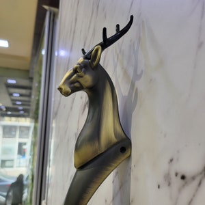 May include: A gold-toned deer head door handle with black antlers. The handle is mounted on a white marble wall.