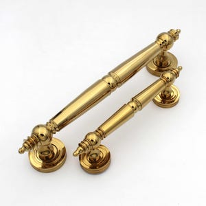 May include: Two gold-colored cabinet handles. One is larger, with a length of approximately 10 inches, and the other is smaller, about 6 inches long. Both have a classic, ornate design with decorative finials and circular base plates.