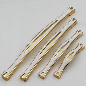 Könnte beinhalten: Ein Satz von fünf Schrankgriffen in verschiedenen Größen. Jeder Griff hat eine goldfarbene Basis mit einer weißen Mitte und goldenen Akzenten. Die Griffe haben ein schlankes, modernes Design, das für Möbel oder Schränke geeignet ist.