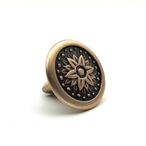 Könnte beinhalten: Ein runder Möbelknopf mit gebürstetem Messing-Finish am äußeren Rand und einer dunklen, vertieften Mitte. Die Mitte zeigt ein erhabenes, achtblättriges Blumenmotiv, das von einem Ring aus kleinen, gleichmäßig verteilten Punkten umgeben ist.