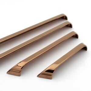 May include: Four bronze-coloured cabinet pulls of varying lengths are arranged diagonally against a white background. Each pull has a sleek, curved design with a slightly angled base, reflecting light.